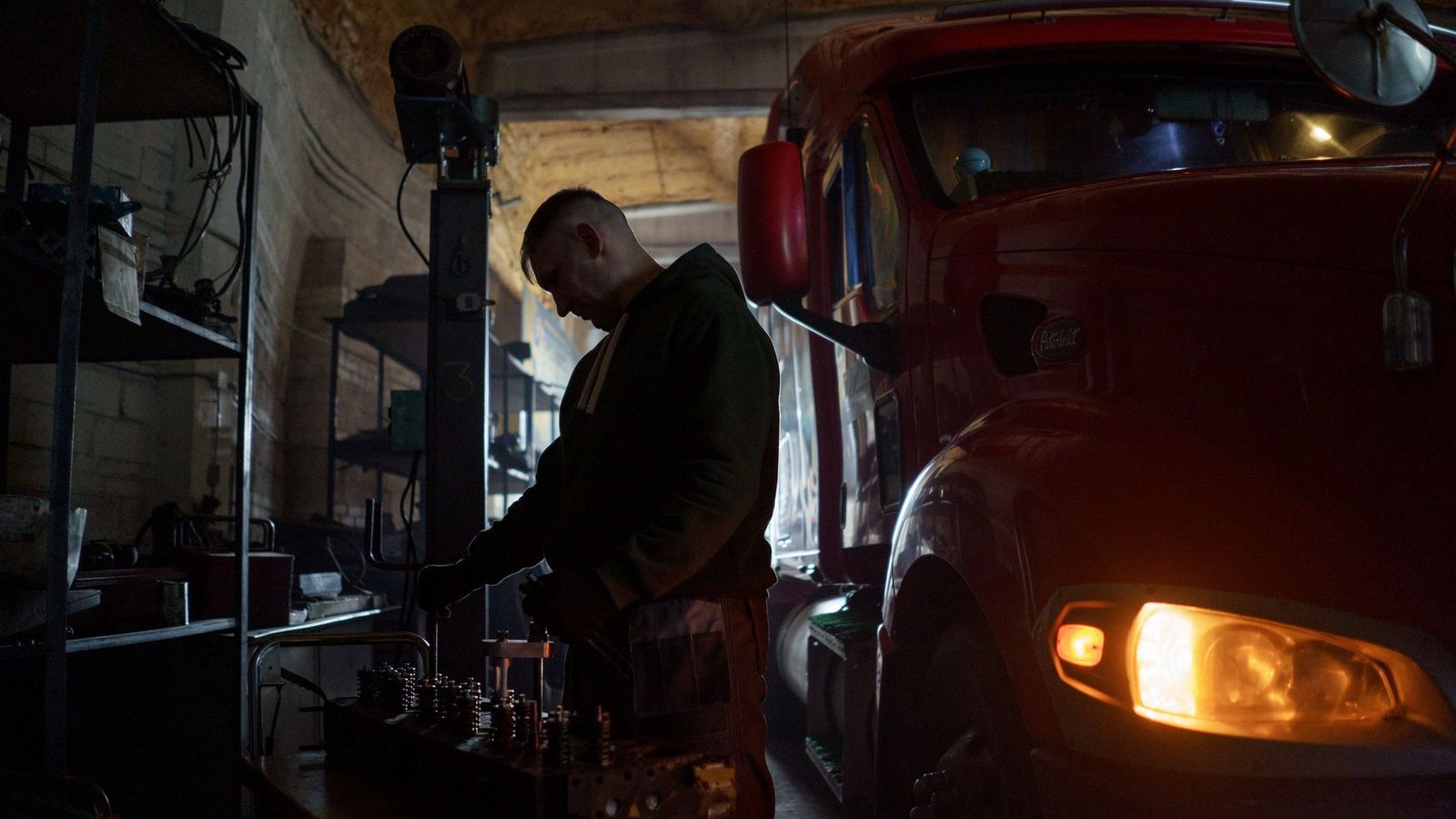 A heavy truck technician working at a bench beside a large red truck in a dim shop, the truck's headlight glowing